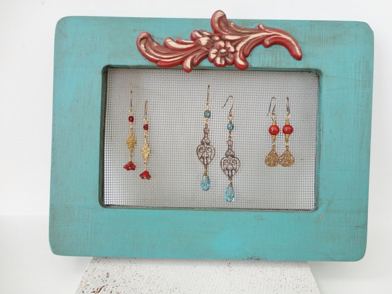 May include: A turquoise blue wooden frame with a wire mesh insert displays three pairs of dangle earrings. The earrings feature gold filigree, red and blue beads.