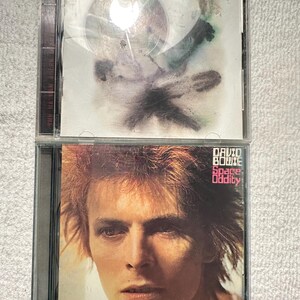 May include: Two David Bowie CD albums. The top album cover features abstract art with the name "David Bowie" at the top. The bottom album cover features a portrait of David Bowie with the title "Space Oddity".