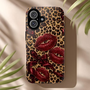 Op de afbeelding: Een smartphonehoesje met een bruin en zwart luipaardpatroon, versierd met drie glanzende rode lippen, een donkerrode satijnen strik en kleine gouden hartjes. Het hoesje heeft een ronde uitsparing voor de camera aan de bovenkant.
