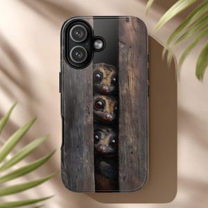 May include: A phone case featuring a detailed image of three wide-eyed lemurs peering through a wooden structure. The case has a glossy finish and a black border, with the camera lenses visible at the top.