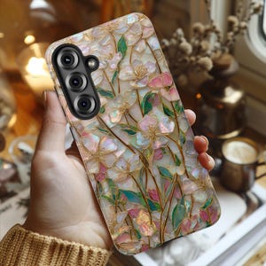 May include: A rectangular smartphone case featuring an iridescent, mother-of-pearl style finish with a floral pattern. The design displays light pink and white blossoms with green leaves and thin gold-colored stems across the entire surface.