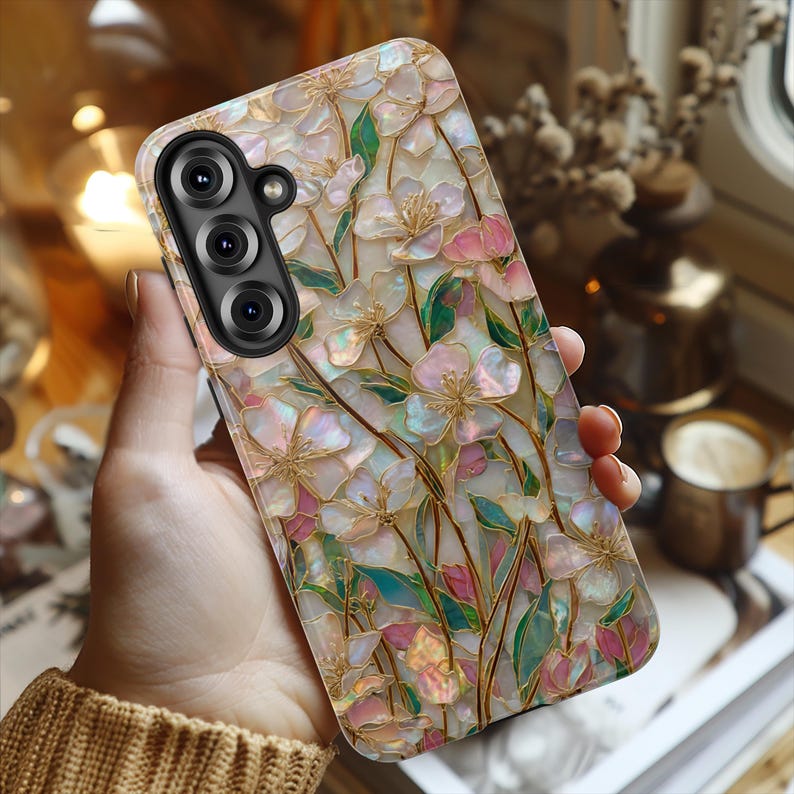 May include: A rectangular smartphone case featuring an iridescent, mother-of-pearl style finish with a floral pattern. The design displays light pink and white blossoms with green leaves and thin gold-colored stems across the entire surface.