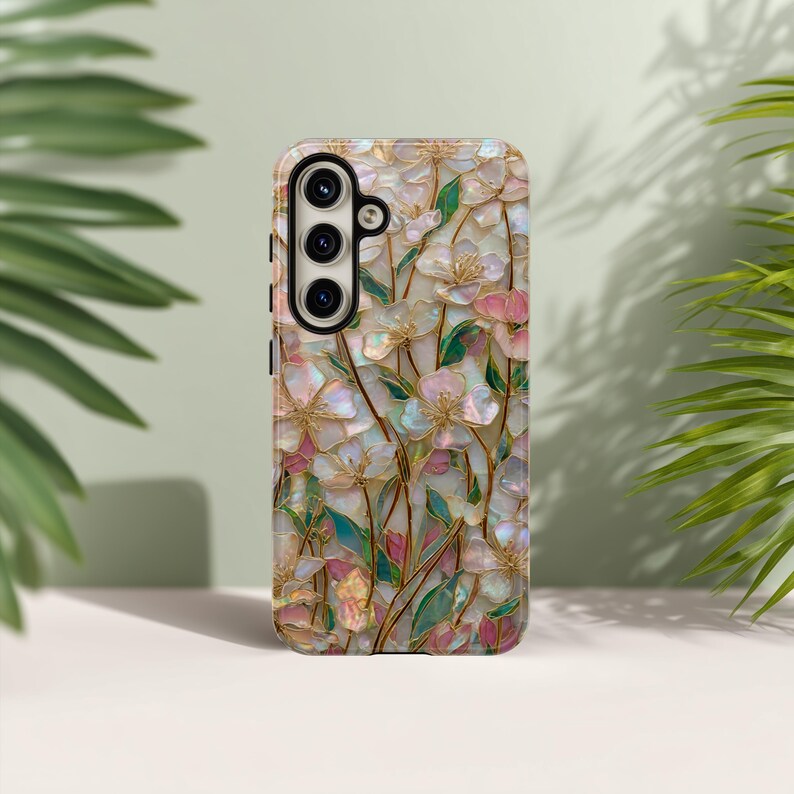 May include: A mobile phone case featuring an iridescent, mother-of-pearl style floral pattern with pink and white petals, green leaves, and thin gold outlines. The case has a black camera module cutout with three circular lens openings on the left.