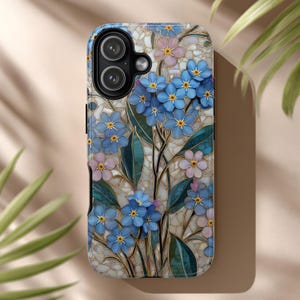 May include: A phone case featuring a floral design with blue and pink flowers, green leaves, and gold accents. The case has a mosaic-like background and a black camera cutout.