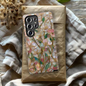 May include: A rectangular smartphone case featuring an iridescent, mother-of-pearl style floral pattern with pink and white petals, green leaves, and gold-toned stems. The case has a black camera module cutout with five circular lens openings.