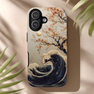 May include: A phone case with a Japanese-inspired design. It features a navy blue wave, a tree with orange blossoms, and a cream background. The case has a dual-camera system and a gold accent.