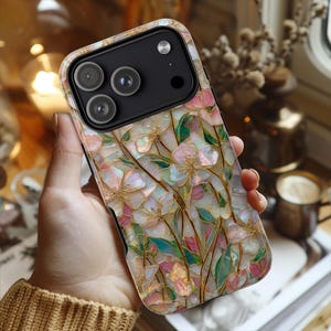 May include: A smartphone case featuring a decorative floral pattern with iridescent, mother-of-pearl style petals in shades of pink, white, and green, accented by thin gold-colored outlines. The case has a black camera module with three lenses.