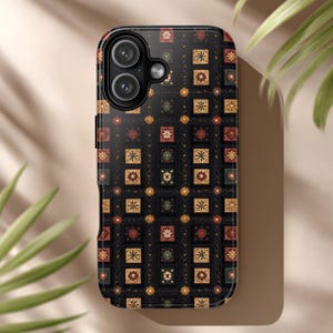 Puede incluir: Funda de teléfono negra con un patrón repetido de cuadrados, flores y formas geométricas en tonos rojos, dorados y verdes. La funda tiene un acabado brillante y un recorte para la cámara.