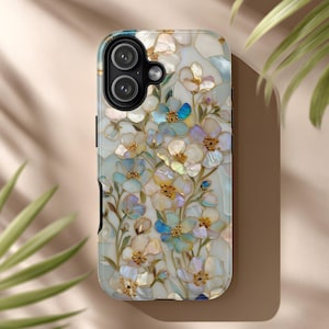 以下が含まれることがあります： 花柄のデザインの携帯電話ケース。ケースは、真珠光沢仕上げの白、青、金の花のパターンが特徴です。ケースには、黒いカメラの切り抜きと黒いサイドボタンが付いています。