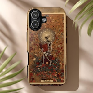 May include: A phone case featuring a tarot card design. The artwork depicts a skeleton reading a book, surrounded by flowers. The case has a gold border and the words "The Reader" at the bottom.