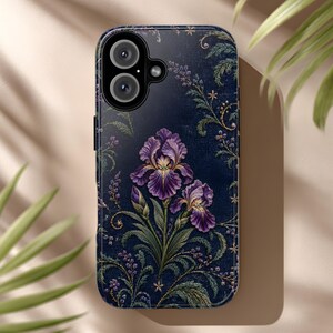 Puede incluir: Una funda de smartphone azul oscuro con un estampado botánico detallado de flores de iris moradas con tallos y hojas verdes. El diseño incluye intrincados patrones de enredaderas y pequeños detalles dorados sobre el fondo azul marino.