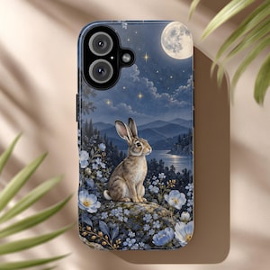 May include: A smartphone case featuring a detailed illustration of a brown rabbit sitting on a rock amidst light blue flowers. The background depicts a serene night landscape with a full moon, twinkling stars, dark mountains, and a calm lake.
