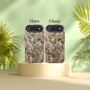 May include: Two mobile phone cases featuring a floral pattern with pink, white, and green petals and gold stems. The left case is labeled Matte and the right case is labeled Glossy. Both cases have a black camera cutout at the top.