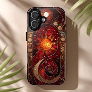 May include: A decorative phone case featuring a stained-glass design with a sun, moon, and star motif. The case has a dark red and gold color scheme with intricate details. The phone case is designed to fit a smartphone.