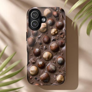 Può includere: Custodia per telefono con design di cioccolato e nocciole. La custodia è marrone scuro con finitura lucida e presenta un motivo di nocciole e pezzi di cioccolato. Le lenti della fotocamera sono visibili nella parte superiore.