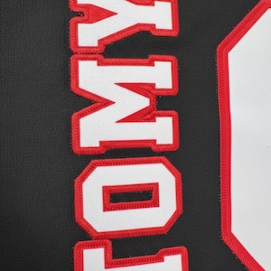 Puede incluir: Primer plano de una tela negra con el nombre "TOMY" bordado en letras blancas con contornos rojos. Las letras est&aacute;n alineadas verticalmente y la tela tiene una apariencia texturizada.