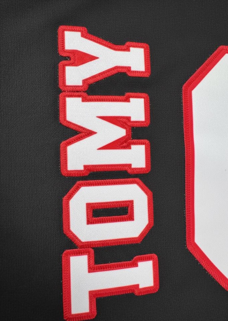 Puede incluir: Primer plano de una tela negra con el nombre "TOMY" bordado en letras blancas con contornos rojos. Las letras est&aacute;n alineadas verticalmente y la tela tiene una apariencia texturizada.