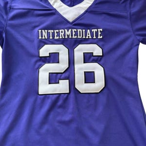 Puede incluir: Una camiseta de f&uacute;tbol morada con detalles blancos y negros. La camiseta muestra la palabra "INTERMEDIATE" y el n&uacute;mero "26" en letras blancas. El n&uacute;mero "6" est&aacute; en el hombro izquierdo y el n&uacute;mero "2" en el hombro derecho.