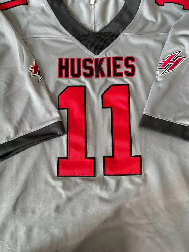 Puede incluir: Camiseta gris con la palabra "HUSKIES" en letras rojas y el n&uacute;mero "11" en rojo. La camiseta tiene detalles negros en el cuello y las mangas, y un logotipo en la manga.