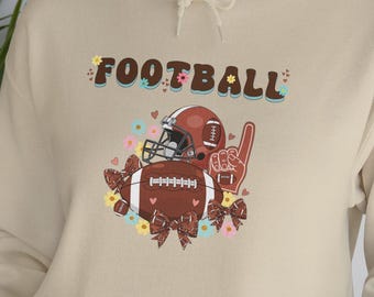 Könnte beinhalten: Ein beigefarbener Kapuzenpullover mit einem Grafikdruck, der den Schriftzug FOOTBALL in braunen Buchstaben zeigt. Das Design umfasst einen braunen Football-Helm, einen Football, einen Schaumstofffinger und braune Schleifen mit kleinen Blumen.