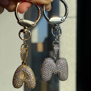 May include: Two keychains with letter pendants. One pendant is the letter "A" in gold with sparkling crystals, and the other is the letter "M" in silver with sparkling crystals. Both keychains have silver-colored clasps.