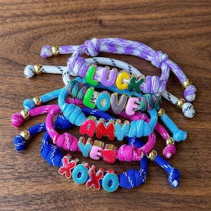 May include: A collection of braided cord bracelets in various colours including purple, blue, pink, and white. Each bracelet features colourful, metallic-finish letter beads spelling out words such as LUCK, LOVE, AMY, YES, and XOXO, with gold-tone beads.