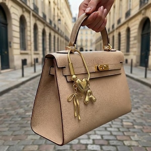 May include: A beige leather handbag with a gold-tone clasp and handle is held up. A gold charm with a heart and other decorative elements hangs from the handle. The bag has a structured design and a classic look.