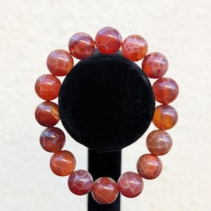 May include: A bracelet featuring large, round, red-orange beads with a marbled appearance. The beads are strung together, forming a circular shape. The bracelet is displayed on a black velvet stand against a white background.