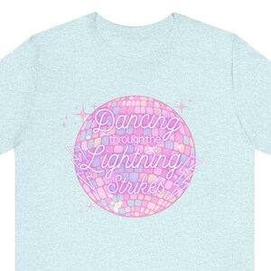 Pode incluir: Camiseta azul clara com um gráfico redondo em estilo mosaico em rosa, roxo e branco. O gráfico apresenta o texto "Dancing through the Lightning Strikes" em uma fonte estilizada, com estrelas.