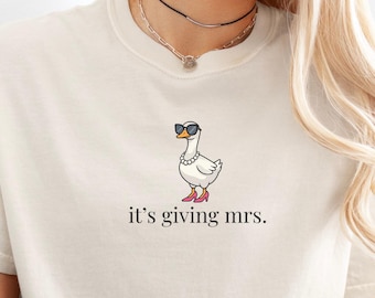 Puede incluir: Camiseta color crema con un dibujo de un ganso con gafas de sol, un collar de perlas y tacones rojos. El texto "it's giving mrs." está impreso debajo. La camiseta es de material suave.