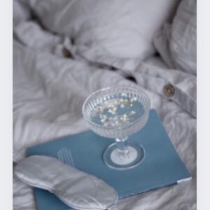 Op de afbeelding: Een heldere champagnecoupe gevuld met vloeistof en kleine witte bloemen staat op een blauw notitieboek. Een zilveren oogmasker ligt op het notitieboek. De items liggen op een bed met wit beddengoed.