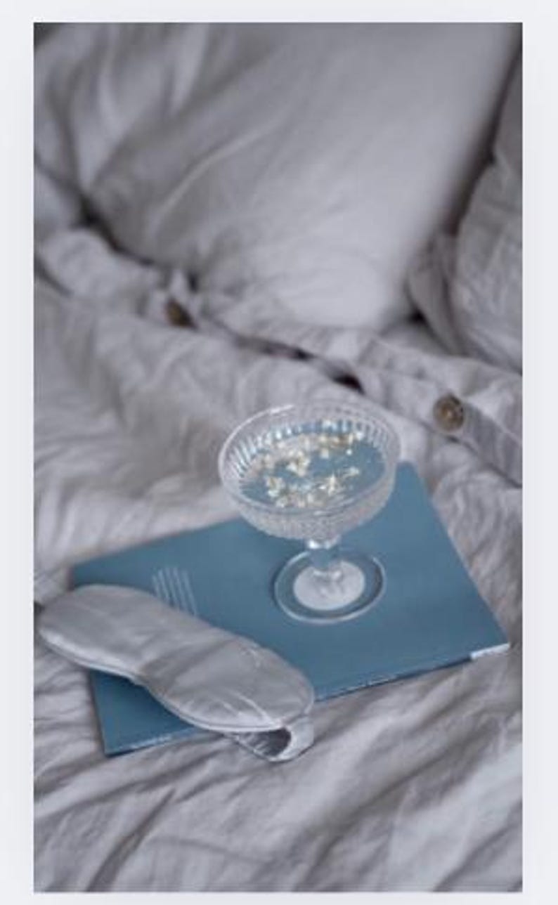 Op de afbeelding: Een heldere champagnecoupe gevuld met vloeistof en kleine witte bloemen staat op een blauw notitieboek. Een zilveren oogmasker ligt op het notitieboek. De items liggen op een bed met wit beddengoed.