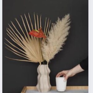 Op de afbeelding: Een beige, golvende vaas bevat gedroogde palmbladeren, pampasgras en een rode anthuriumbloem. Een witte beker staat op een houten kast met een grijze bovenkant. Een hand reikt naar de beker tegen een donkergrijze achtergrond.