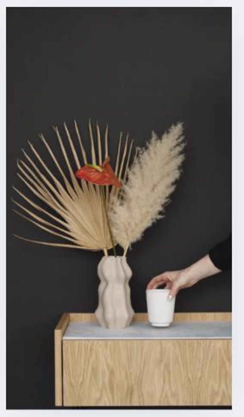 Op de afbeelding: Een beige, golvende vaas bevat gedroogde palmbladeren, pampasgras en een rode anthuriumbloem. Een witte beker staat op een houten kast met een grijze bovenkant. Een hand reikt naar de beker tegen een donkergrijze achtergrond.