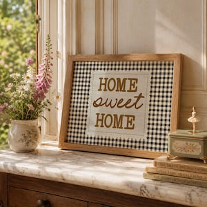 May include: A framed sign with the words "HOME sweet HOME" in gold lettering, set against a black and white checkered background. A small music box and books are on a marble surface, with a vase of flowers to the left.
