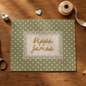 May include: A rectangular sage green fabric panel featuring a white polka dot pattern. Centered is a rectangular cream-colored fabric patch with a delicate lace border, embroidered with the gold-colored cursive text Pippa James.