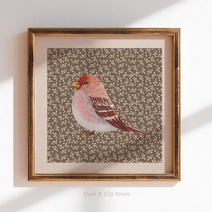 May include: A framed watercolor print of a small bird with a red head, pink chest, and brown wings. The bird is set against a brown background with a delicate floral pattern. The frame is gold. The text "Duck & Lily Press" is at the bottom.