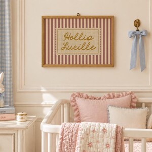 May include: A rectangular wooden frame containing a print with vertical maroon stripes and a central beige scalloped patch featuring the gold cursive text Hollis Lucille. The frame is mounted on a wall above a white crib with pink bedding.