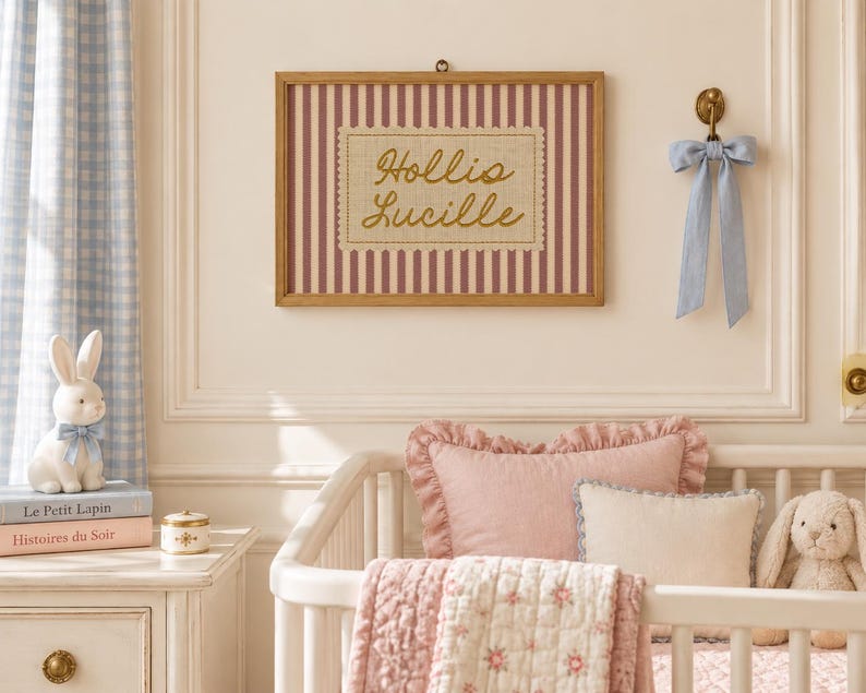 May include: A rectangular wooden frame containing a print with vertical maroon stripes and a central beige scalloped patch featuring the gold cursive text Hollis Lucille. The frame is mounted on a wall above a white crib with pink bedding.