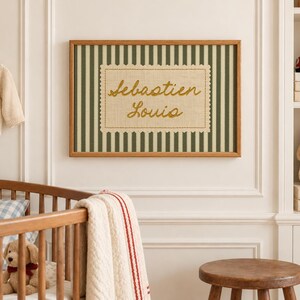 May include: A rectangular wall art piece in a wooden frame featuring a dark green and white vertical striped background. Centered is a beige fabric patch with scalloped edges, embroidered with the name Sebastien Louis in gold thread.