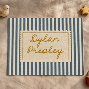 May include: A rectangular fabric sign featuring a blue and white vertical striped pattern. Centered is a beige scalloped-edge patch with the name Dylan Presley embroidered in gold thread. A small wooden bird and a red house are nearby.