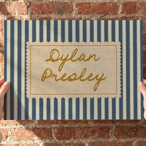 May include: A rectangular fabric sign featuring a blue and white vertical striped pattern. Centered is a beige scalloped-edge patch with the name Dylan Presley embroidered in gold cursive thread, framed by a simple gold stitched border.