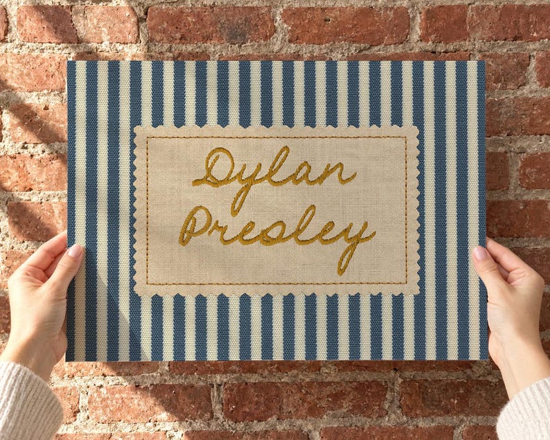 May include: A rectangular fabric sign featuring a blue and white vertical striped pattern. Centered is a beige scalloped-edge patch with the name Dylan Presley embroidered in gold cursive thread, framed by a simple gold stitched border.