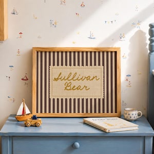 May include: A rectangular wooden frame containing a fabric insert with dark brown vertical stripes and a central cream-colored scalloped label featuring the embroidered gold text Sullivan Bear in a cursive script font.