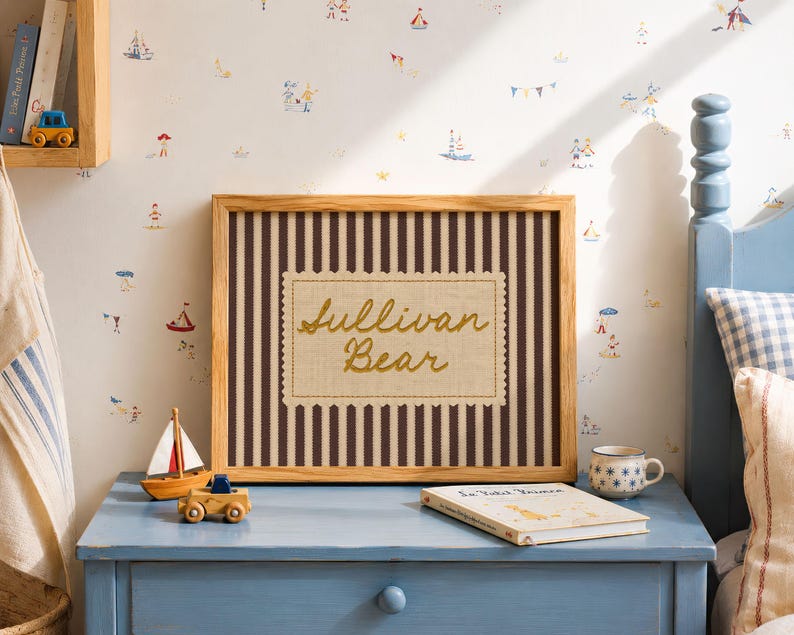 May include: A rectangular wooden frame containing a fabric insert with dark brown vertical stripes and a central cream-colored scalloped label featuring the embroidered gold text Sullivan Bear in a cursive script font.