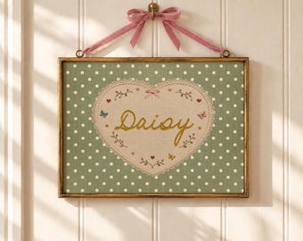 May include: A rectangular framed wall hanging featuring a sage green fabric background with white polka dots. A central cream-colored heart with lace trim displays the embroidered name Daisy in gold, accented by small butterflies, hearts, and a pink bow.