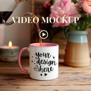 May include: A white coffee mug with a pink handle and rim is being filled with coffee. The mug has the text "your design here" in black script. The image also includes a candle, flowers, and the text "VIDEO MOCKUP".