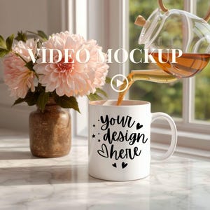May include: A white coffee mug with the text "your design here" in black script, stars, and hearts. Tea is being poured into the mug from a glass teapot. A vase of pink flowers sits to the left of the mug.