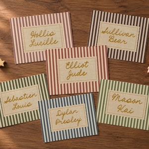 May include: Five rectangular cards with vertical stripes in muted colors, each featuring a central cream-colored label with scalloped edges. The labels display names in gold cursive script: Hollis Lucille, Sullivan Bear, Elliot Jude, Sebastien Louis, Dylan Presley, and Mason Kai. A small wooden star and a miniature rabbit figurine are present.