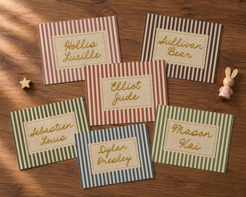 May include: Five rectangular cards with vertical stripes in muted colors, each featuring a central cream-colored label with scalloped edges. The labels display names in gold cursive script: Hollis Lucille, Sullivan Bear, Elliot Jude, Sebastien Louis, Dylan Presley, and Mason Kai. A small wooden star and a miniature rabbit figurine are present.